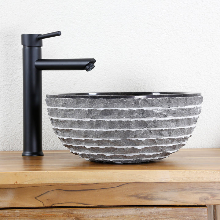 Round countertop washbasin made of natural stone with a structured surface on a wooden top, combined with a black stand-alone tap