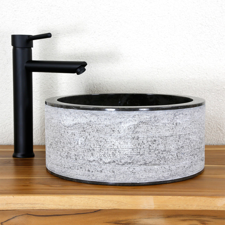 Round countertop washbasin made of natural stone with a striking line structure, combined with a black stand-mounted tap on wood.