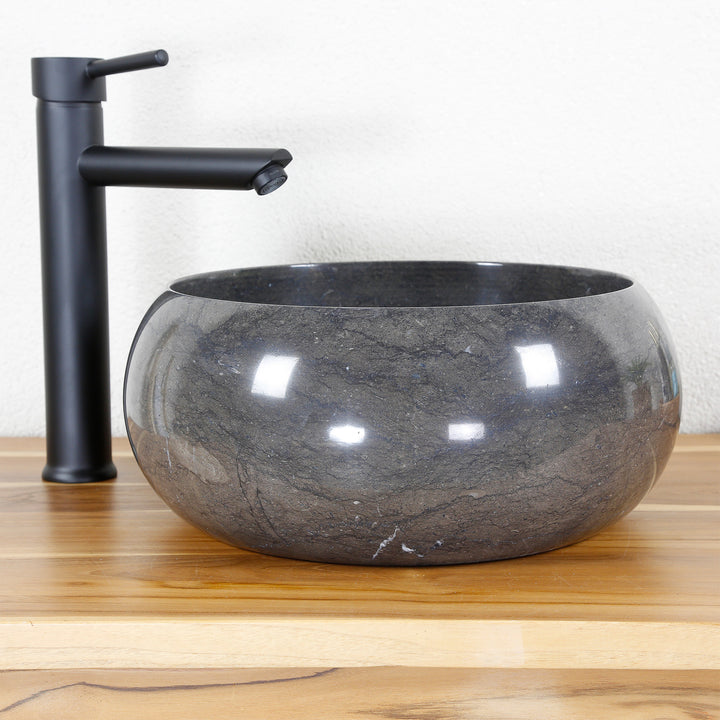 Round stone sink made of marble with dark grain on a wooden top, combined with a black stand-alone tap in front of a light wall
