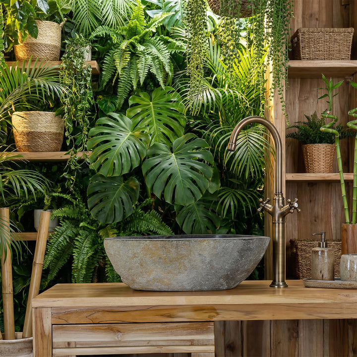 Countertop basin made of river stone in green spa bathroom on wooden vanity unit, combined with brass fittings and lush plant ambience
